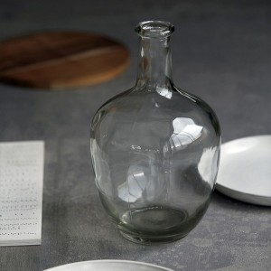 Gros plan sur le vase bouteille en verre transparent posé sur une surface en bois avec assiette - House Doctor - Woodely